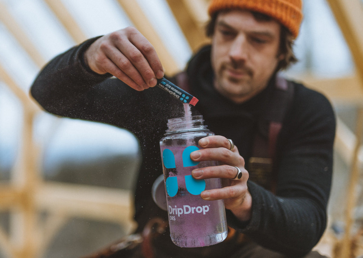 A male young adult pouring DripDrop electrolyte powder into a water bottle with DripDrop's logo outdoors