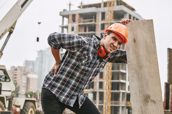 A construction worker wearing a hard hat and plaid shirt grimaces in pain while holding his lower back, leaning against a wooden plank at a construction site.