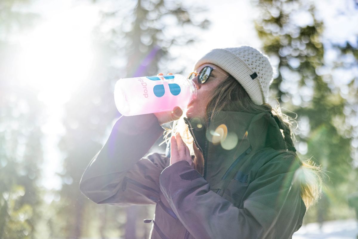 A woman in a winter coat and beanie drinks from a DripDrop bottle outdoors, with sunlight streaming through the trees, highlighting the cold weather setting.