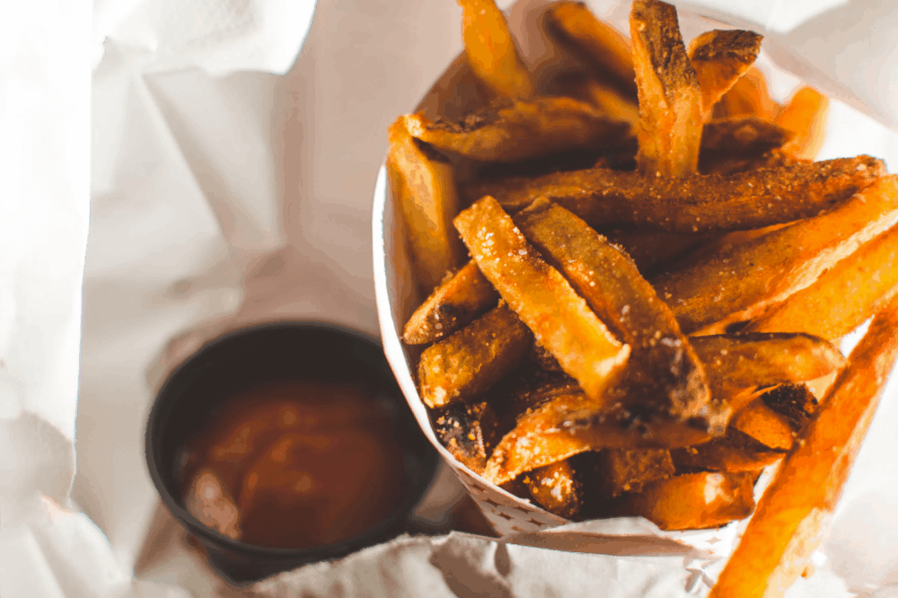 A close-up of crispy golden French fries served in a paper cone with a side of ketchup, highlighting their texture and seasoning.