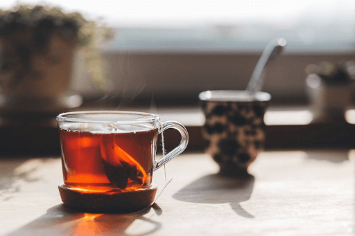 A steaming cup of tea sits on a wooden table, with a blurred mug in the background.