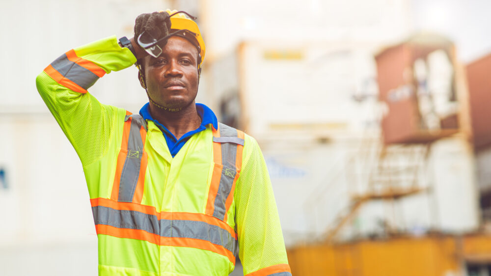 A construction worker in a high-visibility uniform wipes sweat from his forehead, appearing fatigued from the heat.