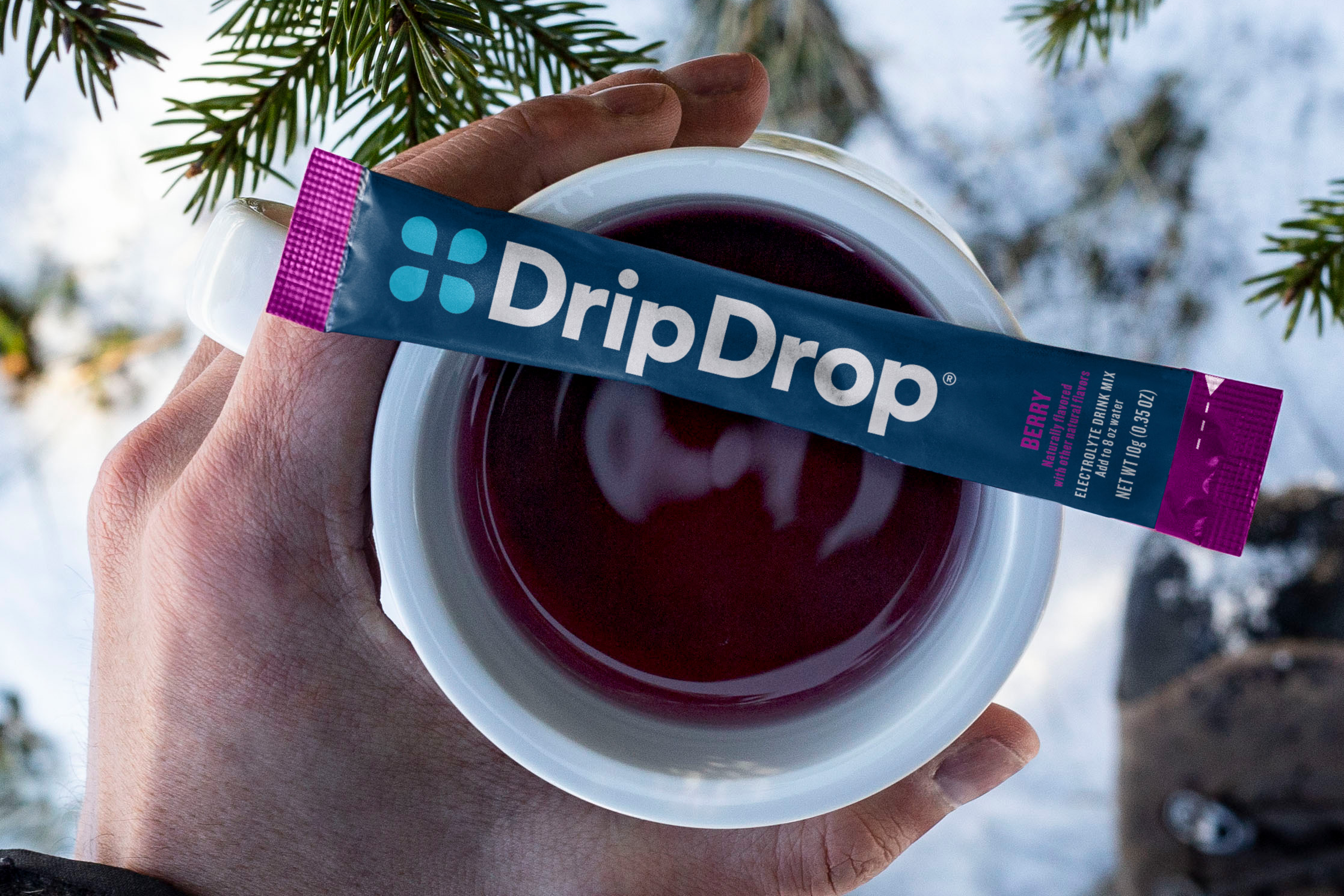A hand holding a mug with warm liquid and a DripDrop stick with snow in the background