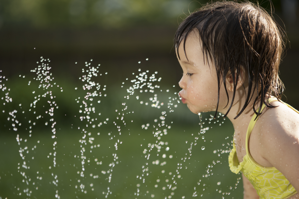 A toddler in a yellow swimsuit plays with a sprinkler, staying cool.