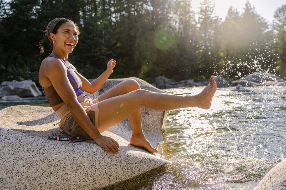 A woman in athletic wear sits on a rock by a river, splashing water with her foot and smiling in the sunlight. DripDrop hydration packets rest beside her.