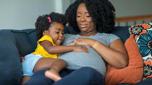 A pregnant mother relaxes on the couch while her young daughter gently touches her belly