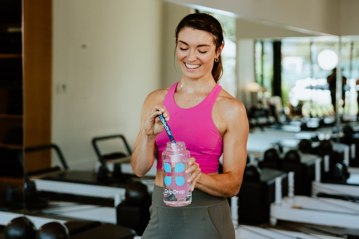 Woman in a pink top standing in a pilates studio pouring a stick of DripDrop into a water bottle
