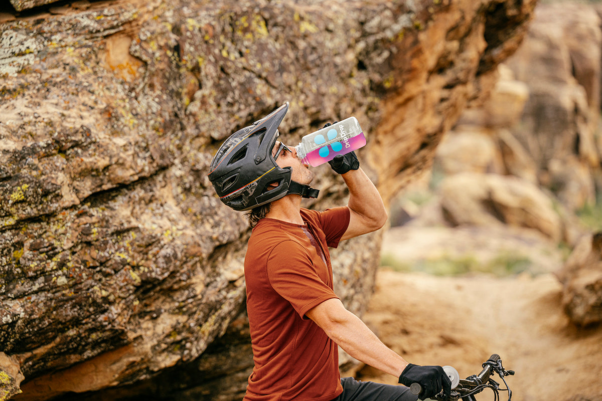 Mountain biker in helmet drinking from a bottle while resting on rocky terrain.