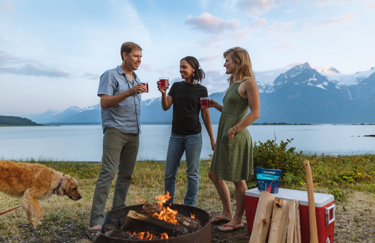 Three friends toast with red drinks by a lakeside campfire, with DripDrop packets on a red cooler and mountains in the background.