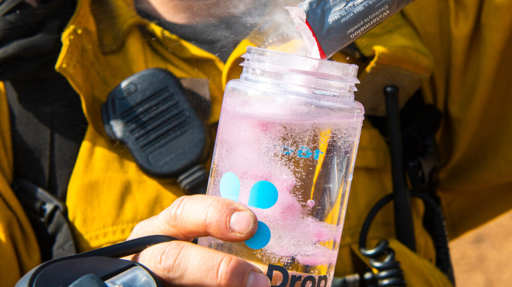A person in a yellow uniform pouring an electrolyte powder into a water bottle.