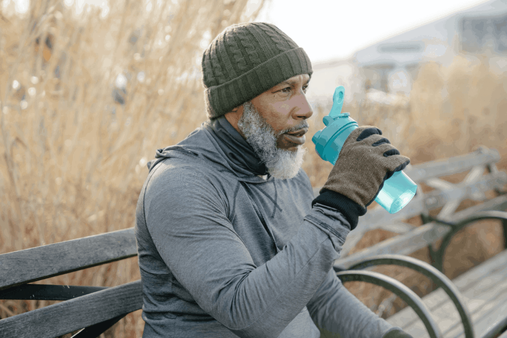 Senior man in winter clothing sitting on a bench, drinking from a blue water bottle to stay hydrated.