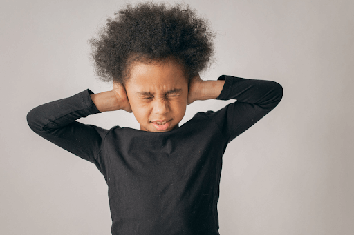 Young child in a black long-sleeve shirt pressing their hands against their ears with a pained expression, eyes closed.