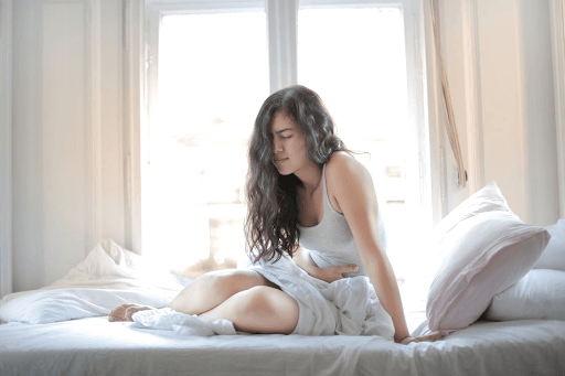 A woman sits on her bed, clutching her stomach in discomfort