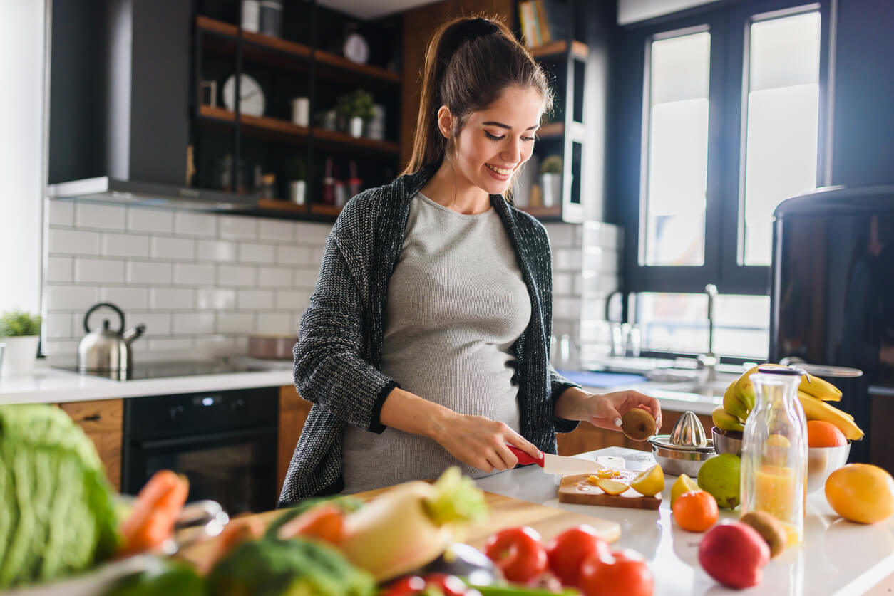 Smiling pregnant woman preparing fresh fruit in a modern kitchen, surrounded by healthy ingredients.