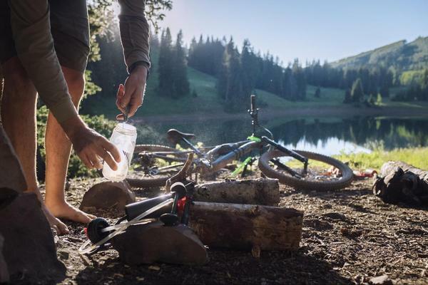 A camper refills a water bottle near a mountain lake, with bicycles resting nearby, highlighting outdoor hydration and adventure in a scenic wilderness setting.