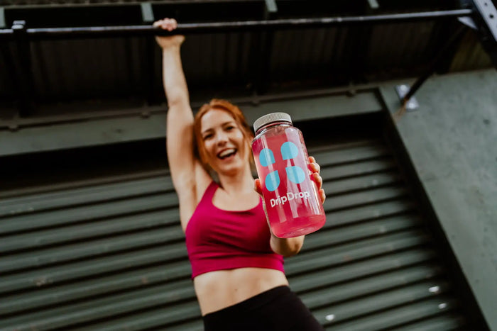 A redheaded woman standing in front of a garage holding a bottle with the DripDrop logo filled with pink liquid in one hand and holding onto a bar with the other