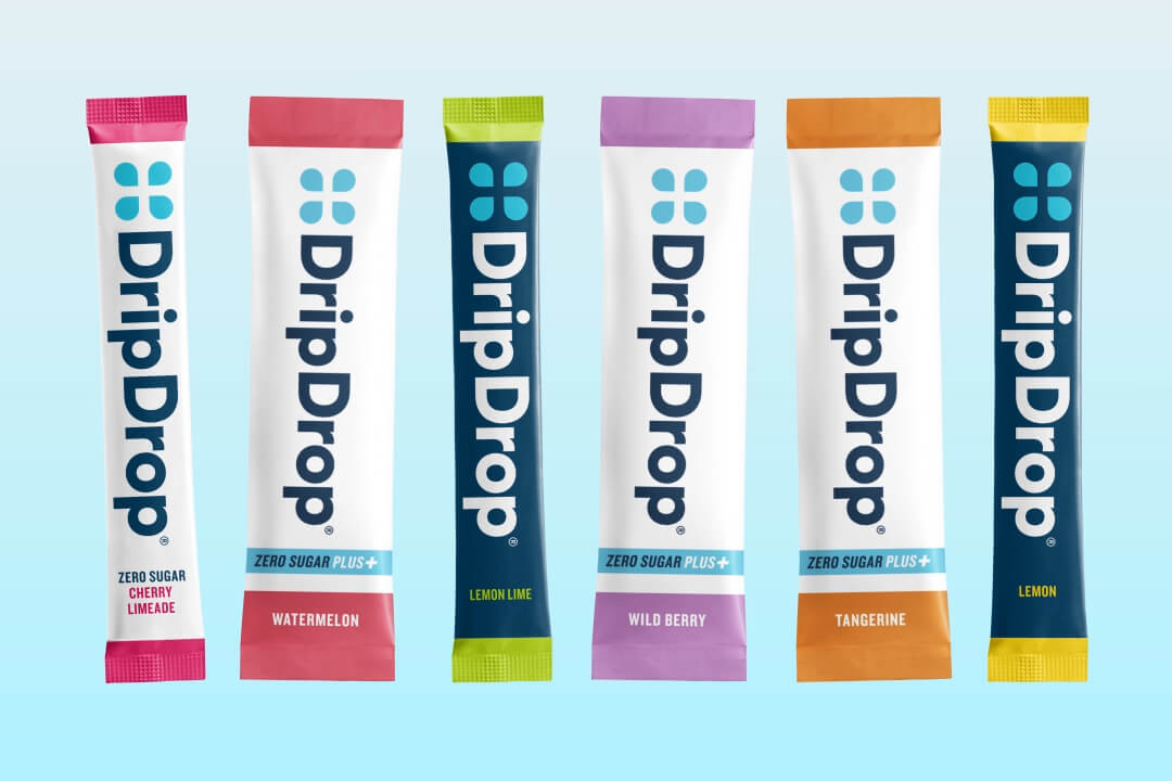 Six DripDrop packs arranged upright in a row on a light blue background. The packs feature different flavor colors: cherry limeade, watermelon, lemon lime, wild berry, orange tangerine, and yellow lemon.