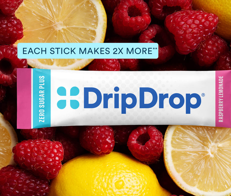 A DripDrop Zero Sugar Plus raspberry lemonade stick pack placed over a close-up background of fresh raspberries and lemon slices. The package is white with blue branding and pink side accents, with a light blue text banner above reading 'EACH STICK MAKES 2X MORE'.