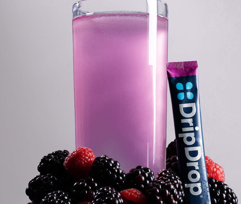 A glass of water with pink liquid surrounded by berries with a DripDrop stick leaning against it