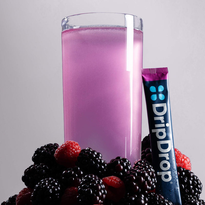 A glass of water with pink liquid surrounded by berries with a DripDrop stick leaning against it