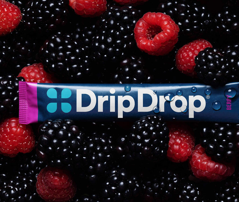 A DripDrop berry-flavored stick product wrapped in a blue and pink package, laid horizontally across a dense pile of fresh blackberries and raspberries.