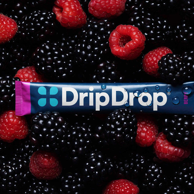 A DripDrop berry-flavored stick product wrapped in a blue and pink package, laid horizontally across a dense pile of fresh blackberries and raspberries.