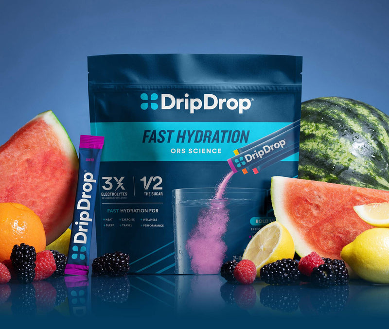 A stand-up pouch of DripDrop Fast Hydration ORS powder with a single stick pack in front, surrounded by watermelon slices, lemons, blackberries, raspberries, and an orange on a blue studio background. A glass and pink powder pour are shown in the center.