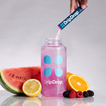 A clear plastic bottle filled with pink liquid and branded DripDrop, with a hand pouring more DripDrop powder from a sachet into the bottle. Watermelon, lemon, orange, and berries are arranged around the bottle on a light gray reflective surface.