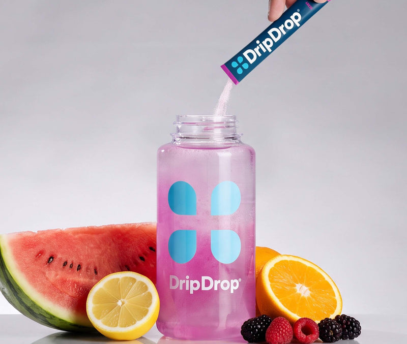 A clear plastic bottle filled with pink liquid and branded DripDrop, with a hand pouring more DripDrop powder from a sachet into the bottle. Watermelon, lemon, orange, and berries are arranged around the bottle on a light gray reflective surface.
