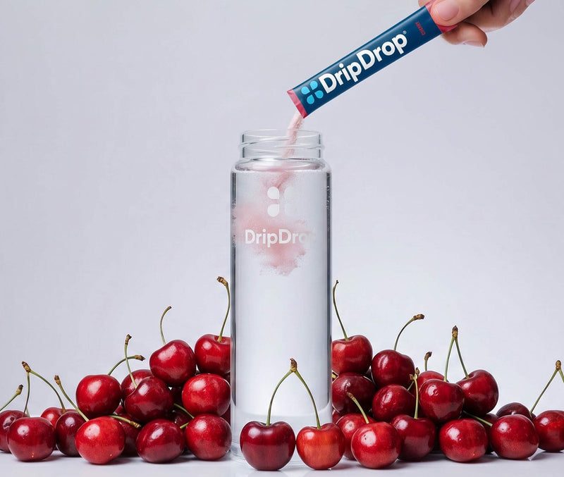 A hand pours a pink liquid from a DripDrop stick packet into a clear bottle, surrounded by fresh red cherries on a light gray background.