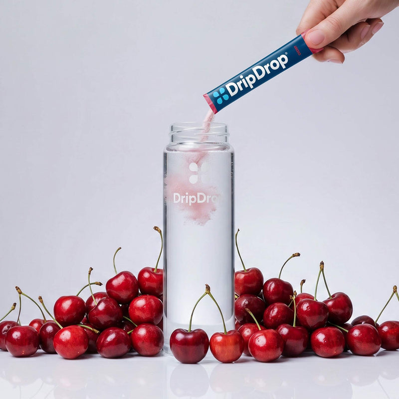 A hand pours a pink liquid from a DripDrop stick packet into a clear bottle, surrounded by fresh red cherries on a light gray background.