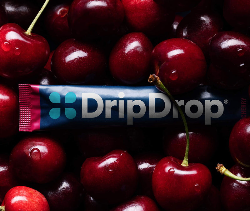 A DripDrop cherry-flavored liquid supplement tube placed horizontally across a dense pile of fresh red cherries. The tube is dark blue with a pink and teal logo and the brand name 'DripDrop' in white.