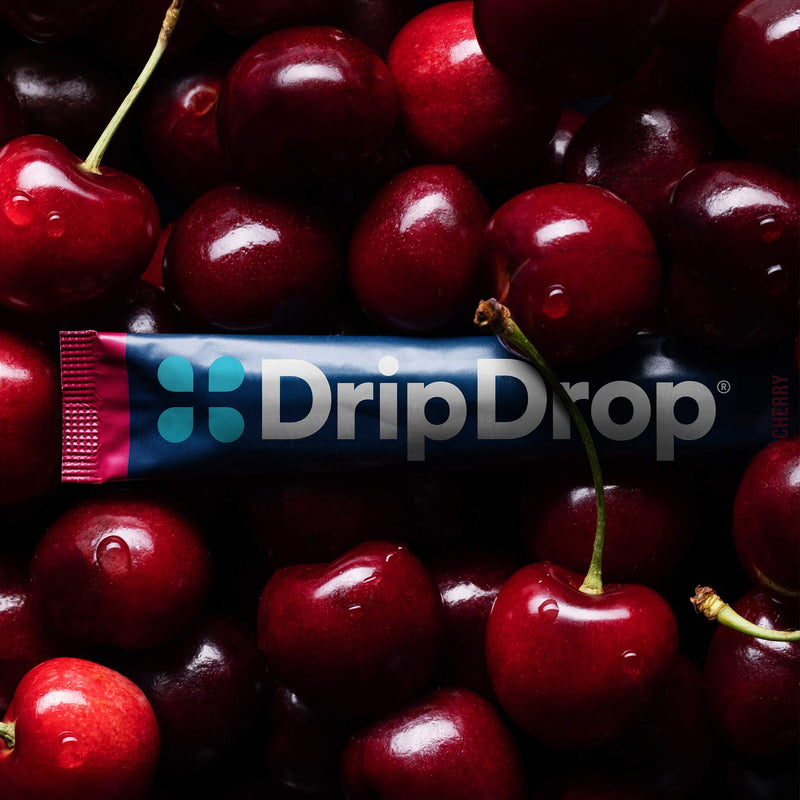 A DripDrop cherry-flavored liquid supplement tube placed horizontally across a dense pile of fresh red cherries. The tube is dark blue with a pink and teal logo and the brand name 'DripDrop' in white.