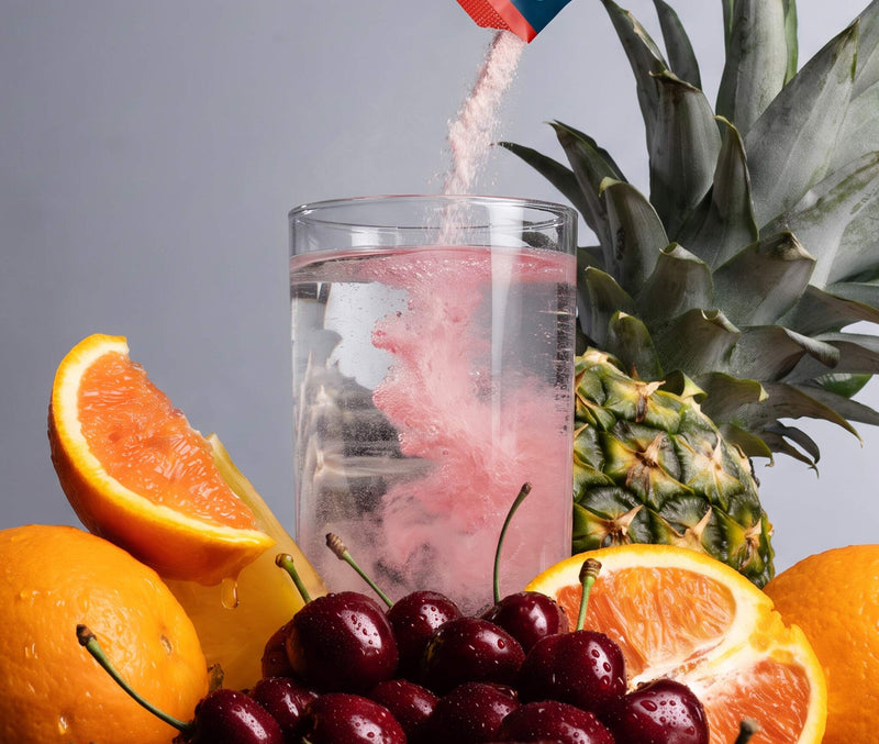 A clear glass of water with pink powder swirling inside, surrounded by fresh fruit including a pineapple, oranges, grapefruits, and dark red cherries. A DripDrop powder packet is pouring the pink powder into the glass against a neutral gray background.