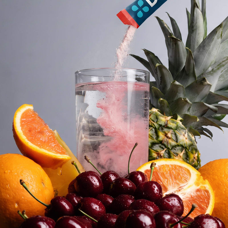 A clear glass of water with pink powder swirling inside, surrounded by fresh fruit including a pineapple, oranges, grapefruits, and dark red cherries. A DripDrop powder packet is pouring the pink powder into the glass against a neutral gray background.