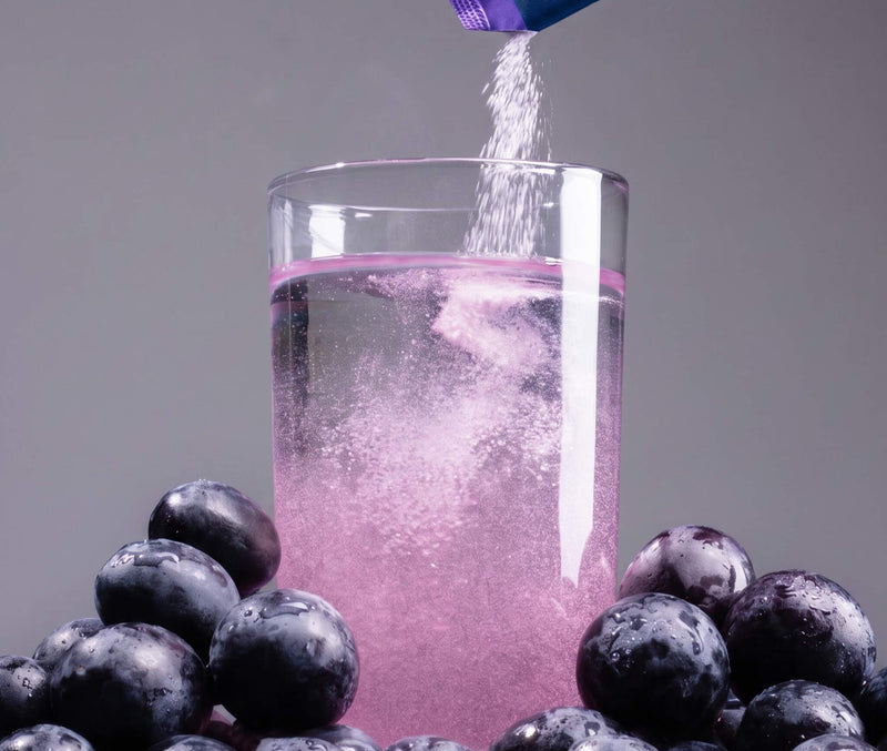 Grape DripDrop stick being poured into a water glass surrounded by grapes on a gray background