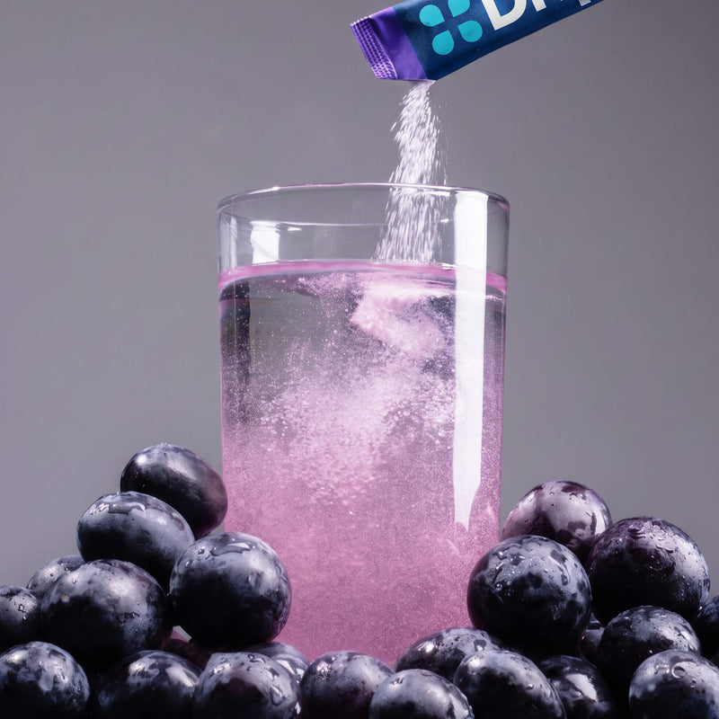 Grape DripDrop stick being poured into a water glass surrounded by grapes on a gray background