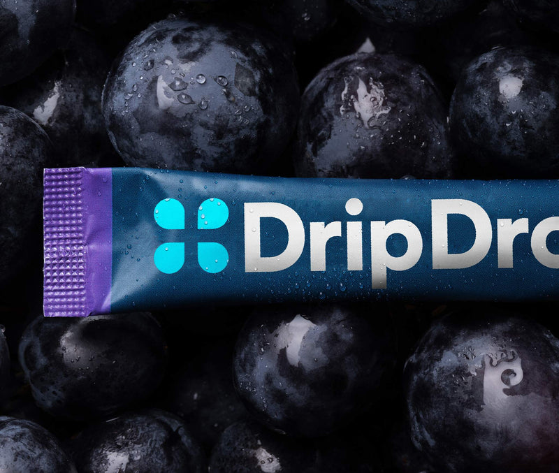 A DripDrop juice pouch lies horizontally across a close-up pile of dark red grapes with water droplets on them. The pouch is dark blue with a purple end cap and the DripDrop logo in bright blue and white.