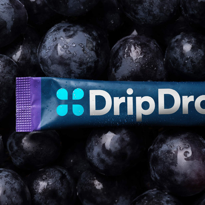 A DripDrop juice pouch lies horizontally across a close-up pile of dark red grapes with water droplets on them. The pouch is dark blue with a purple end cap and the DripDrop logo in bright blue and white.
