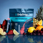 A DripDrop Fast Hydration powder pouch and matching stick pack displayed on a reflective surface with fruit props including strawberries, lemons, oranges, cherries, and pineapple against a blue studio background.