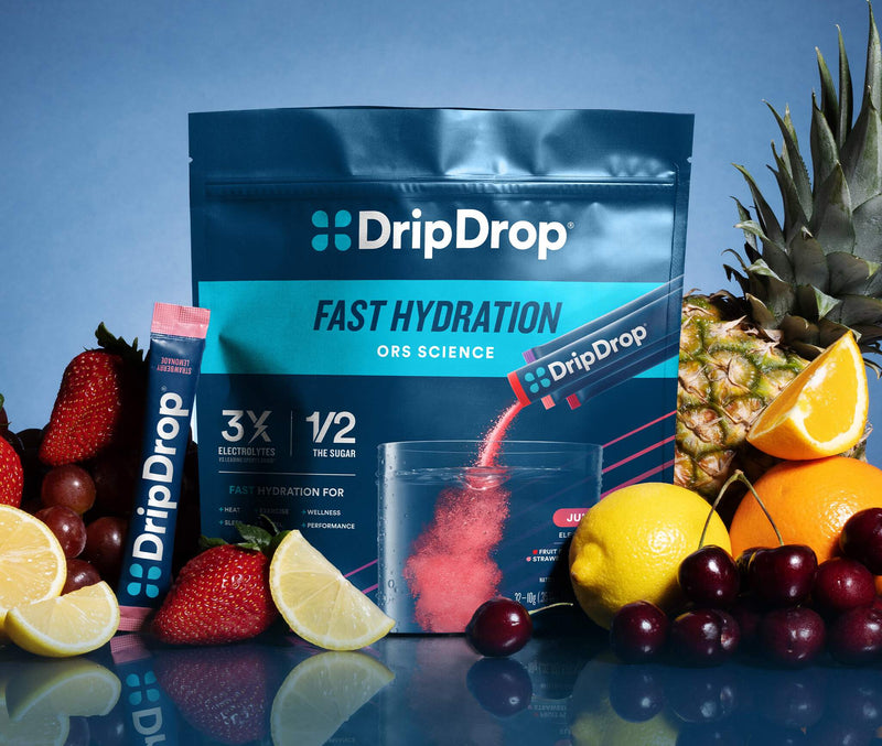 A DripDrop Fast Hydration powder pouch and matching stick pack displayed on a reflective surface with fruit props including strawberries, lemons, oranges, cherries, and pineapple against a blue studio background.