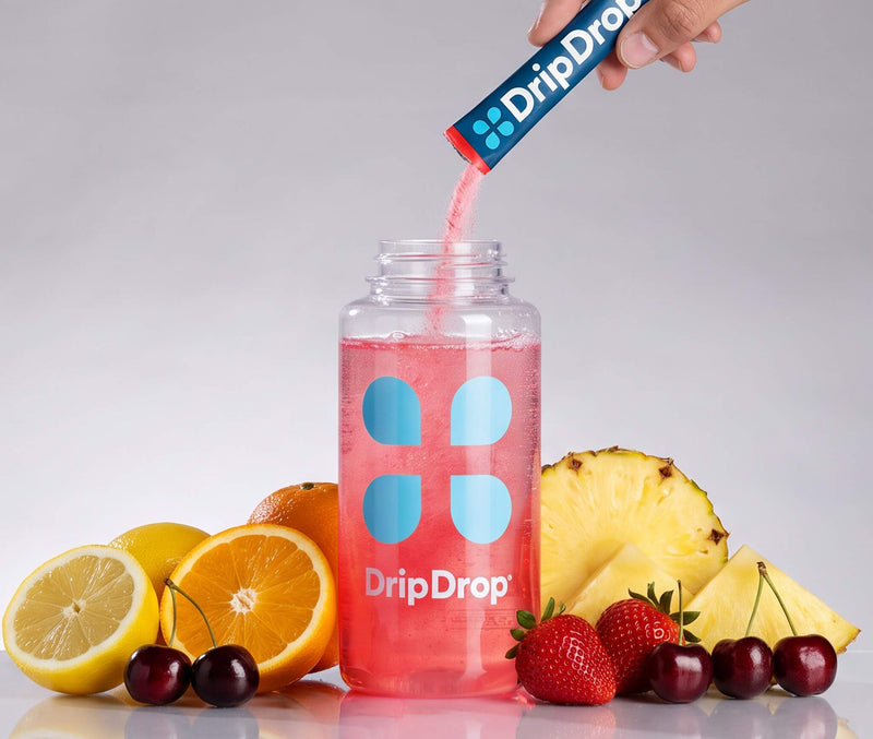 A hand pours pink liquid from a blue DripDrop tube into a clear DripDrop bottle filled with pink drink, surrounded by fruit slices and cherries on a light gray background.