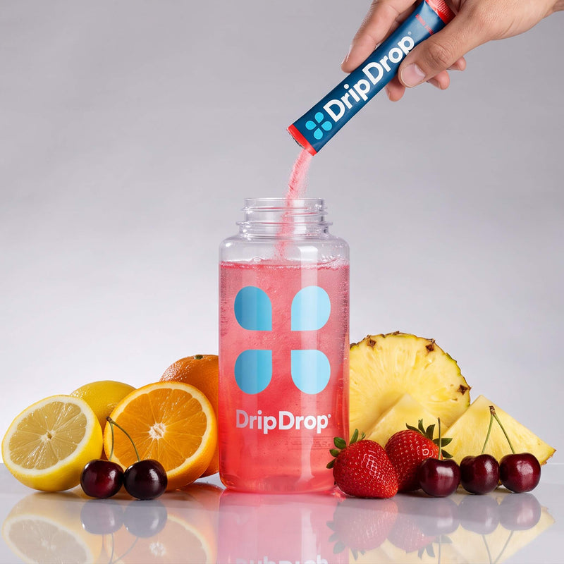 A hand pours pink liquid from a blue DripDrop tube into a clear DripDrop bottle filled with pink drink, surrounded by fruit slices and cherries on a light gray background.