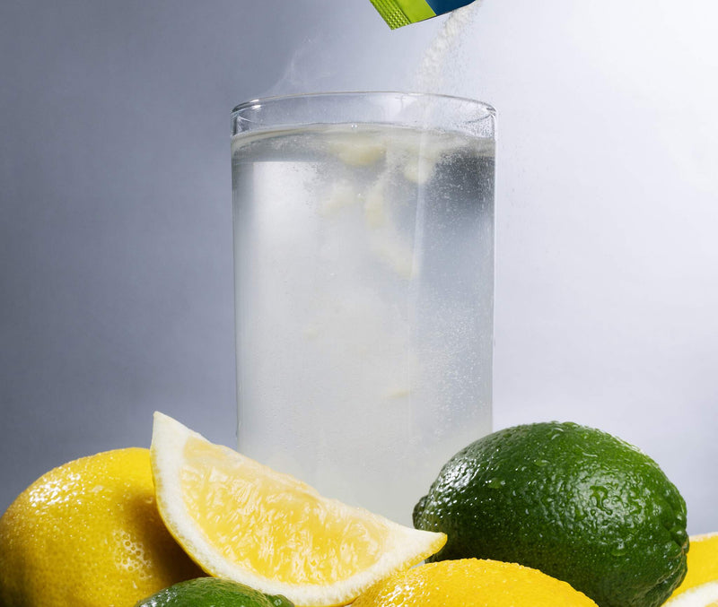 A clear glass of water with lemon and lime slices in the foreground, with a blue and green packet pouring a white powder into the glass against a gray studio background.