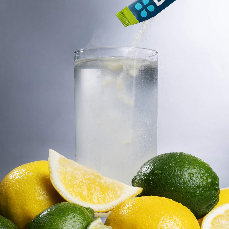 A clear glass of water with lemon and lime slices in the foreground, with a blue and green packet pouring a white powder into the glass against a gray studio background.