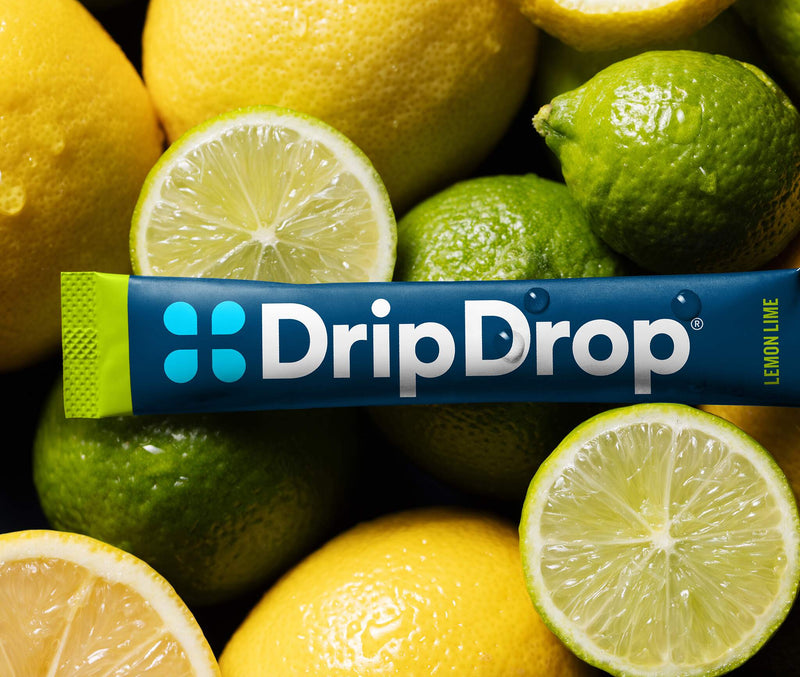 A DripDrop lemon lime flavored stick pack placed horizontally across a pile of whole and sliced lemons and limes. The packaging is dark blue with a green end cap and white brand text.