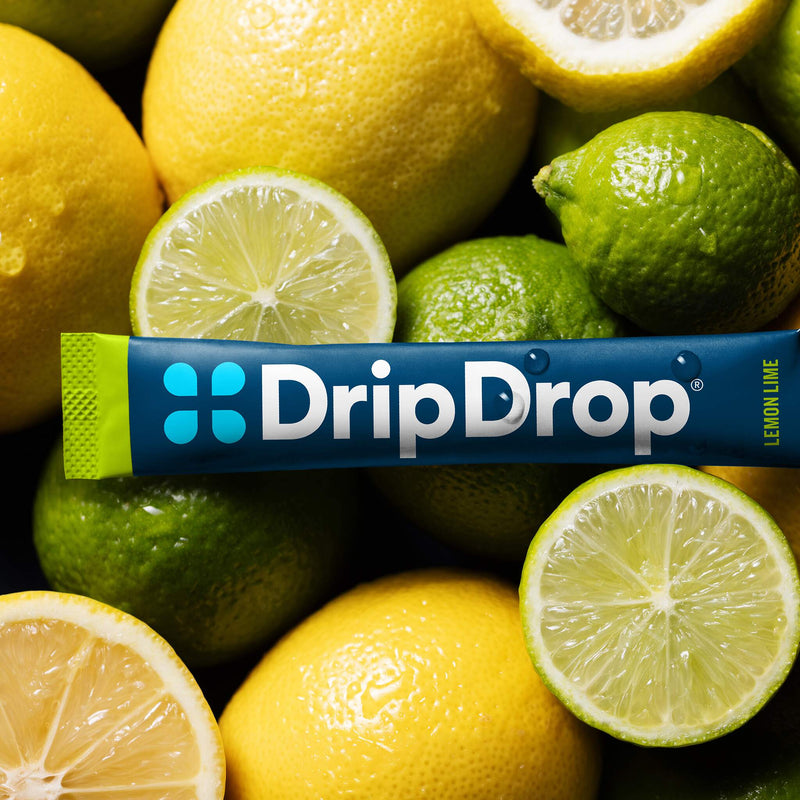A DripDrop lemon lime flavored stick pack placed horizontally across a pile of whole and sliced lemons and limes. The packaging is dark blue with a green end cap and white brand text.