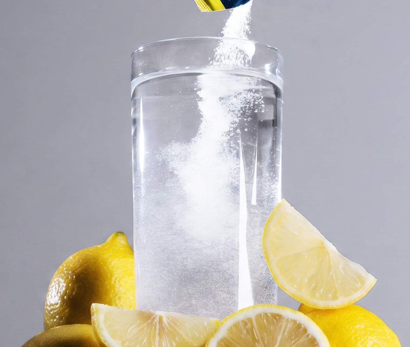 A clear glass filled with water, with a white powder being poured into it from a blue and yellow packet. The glass is surrounded by fresh yellow lemons on a neutral gray background.