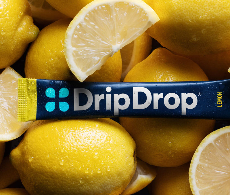 A DripDrop lemon-flavored stick laid horizontally across a pile of bright yellow lemons with sliced wedges and visible water droplets.