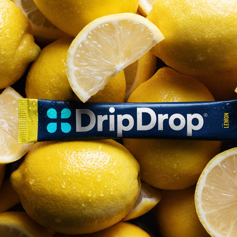 A DripDrop lemon-flavored stick laid horizontally across a pile of bright yellow lemons with sliced wedges and visible water droplets.